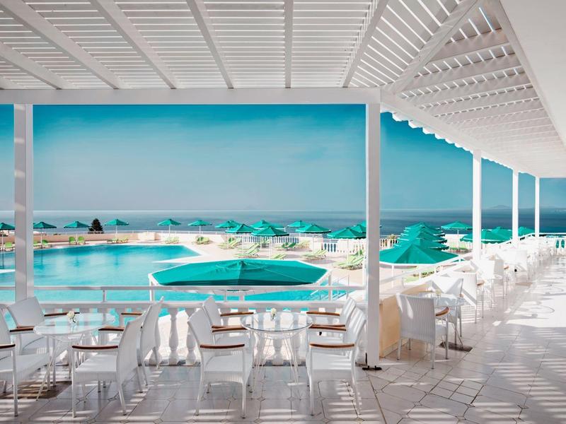 Espace piscine ouvert avec mobilier blanc et parasols turquoise face à la mer.