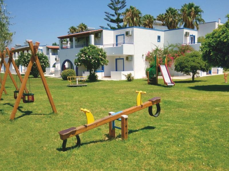 Spielplatz mit Schaukel, Wippe und Rutsche auf grünem Rasen vor weißen Häusern und Palmen.