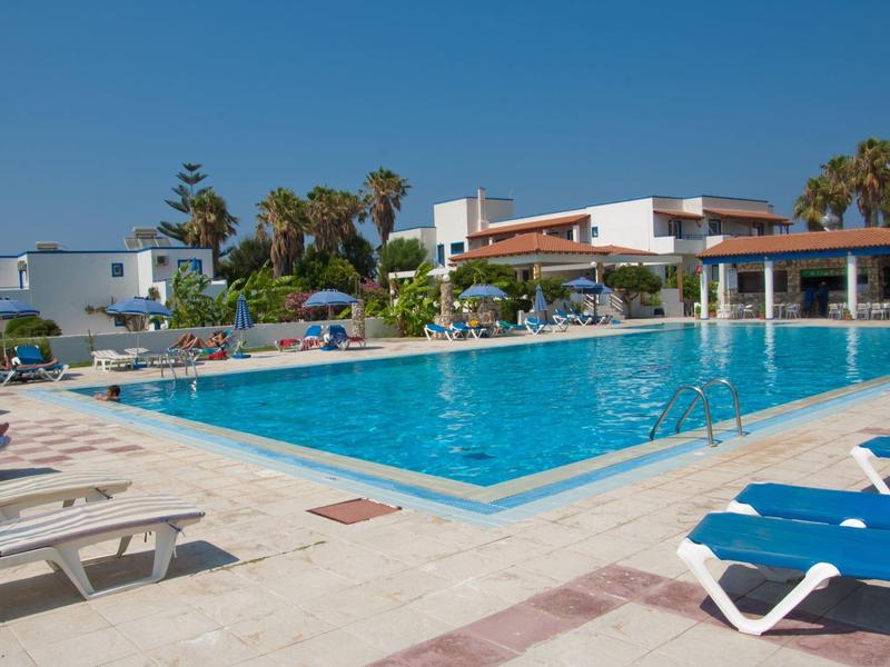 Großer Außenpool mit blauer Wasseroberfläche, weißen Liegestühlen und mehreren Gebäuden im Hintergrund.