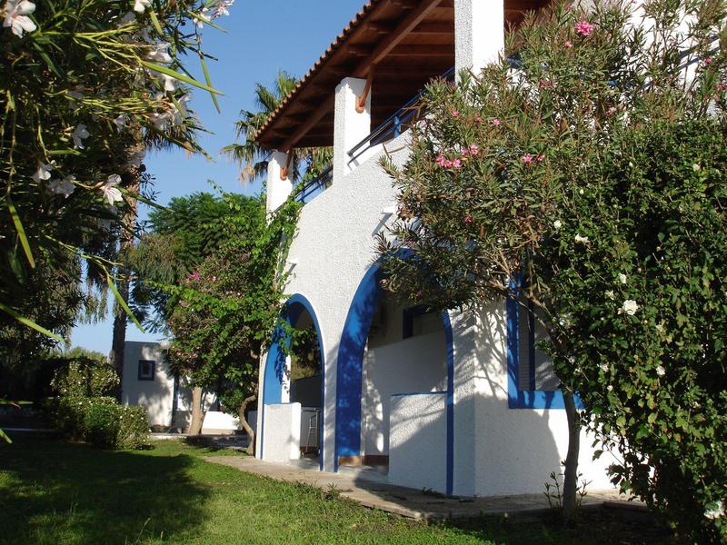 Weißes Haus mit blauem Türbogen, umgeben von grünen Bäumen und Sträuchern bei Sonnenschein.