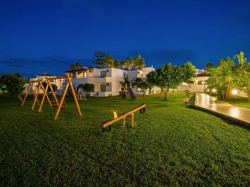 Kinderspielplatz mit Schaukel und Wippe auf grünem Rasen vor beleuchteten Gebäuden bei Nacht.
