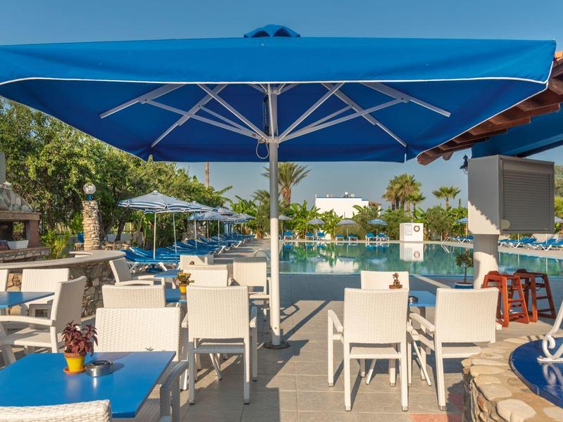 Freizeitbereich mit blauem Sonnenschirm, weißen Stühlen und Tischen, Palmen und Pool im Hintergrund.