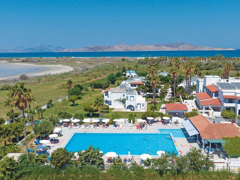 Hotelanlage mit großem blauen Pool, weißen Gebäuden, Palmen und Meer im Hintergrund.