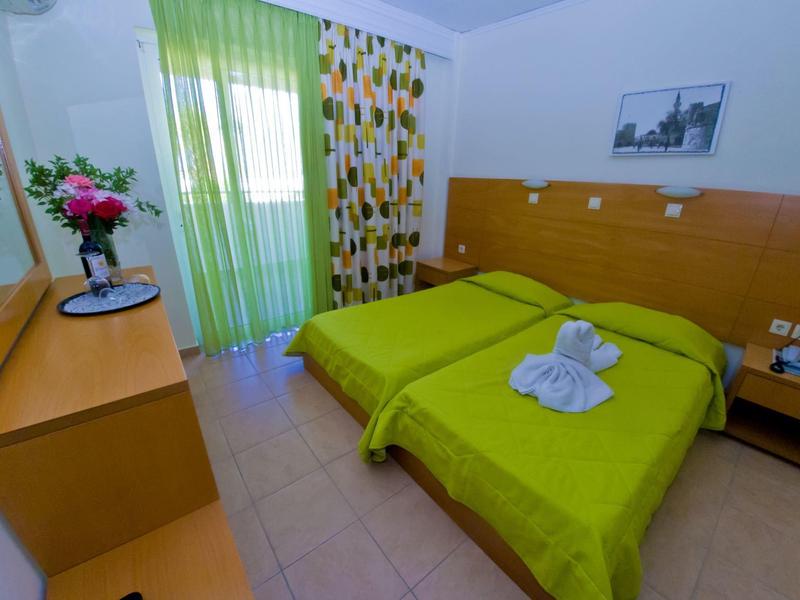 Hell eingerichtetes Schlafzimmer mit zwei grünen Einzelbetten, Holzmöbeln und buntem Vorhang.