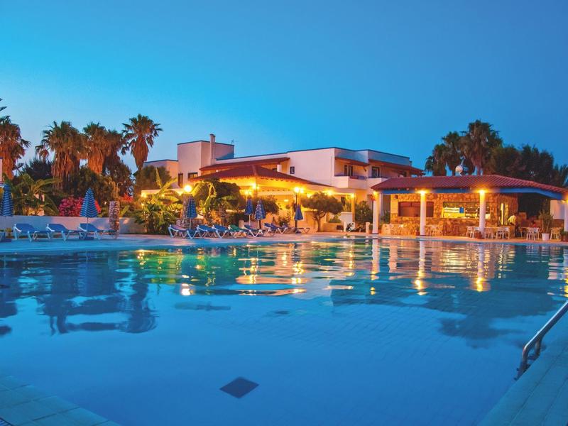 Großer beleuchteter Pool vor modernem Gebäude, umgeben von Palmen und Liegen bei Dämmerung.