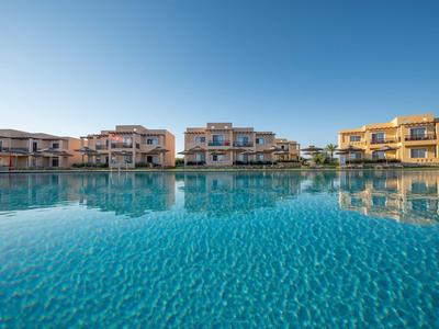 Case vacanze moderne si riflettono in una grande piscina limpida sotto un cielo azzurro.