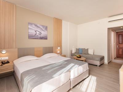 Camera d'hotel luminosa con letto matrimoniale grande, divano grigio e arredamento minimalista.