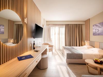 Luminoso camera d'hotel con due letti singoli, grandi tende e parete in legno con specchio rotondo.