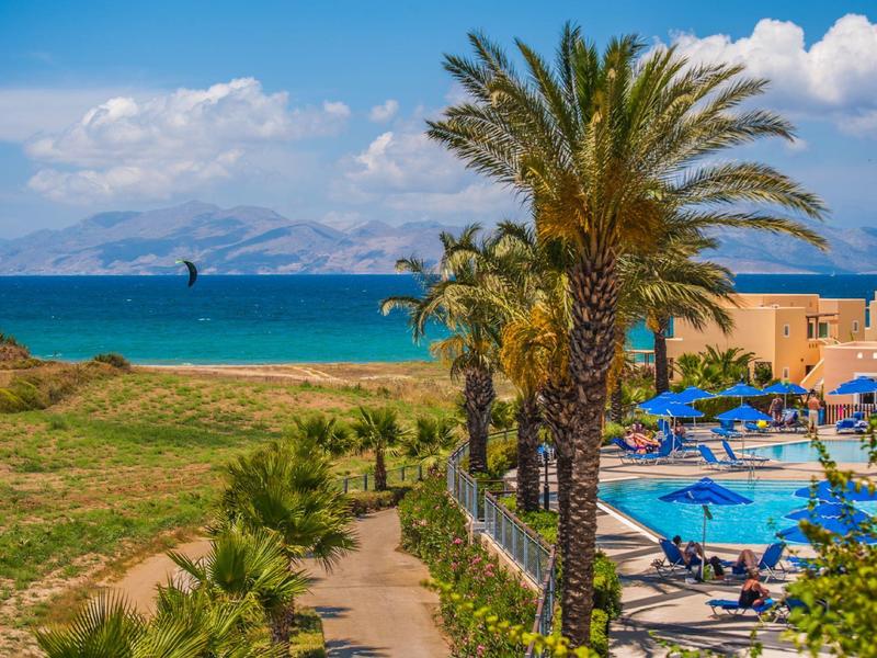 Vista di una piscina con palme, lettini blu e il mare sullo sfondo sotto un cielo sereno.