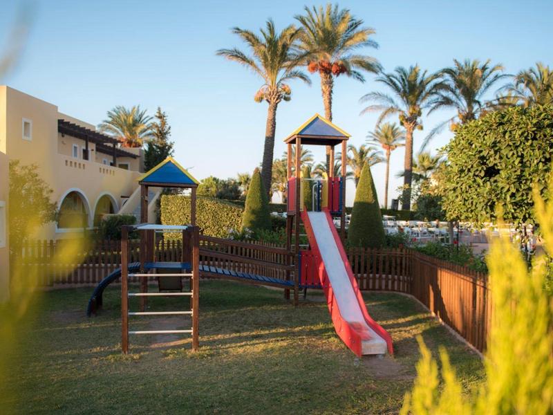 Parco giochi con scivolo e struttura per arrampicarsi in un hotel con palme e prato.