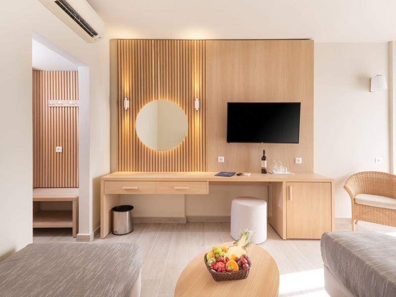 Stanza d'hotel luminosa con due letti, pannello in legno con specchio rotondo e TV, mobili e sedia in legno.