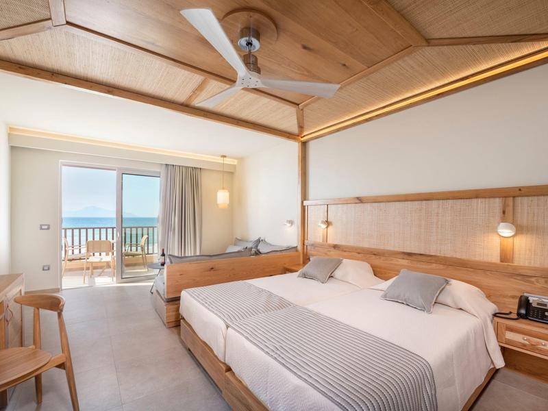 Camera d'hotel luminosa con letto matrimoniale, soffitto in legno e balcone con vista mare.