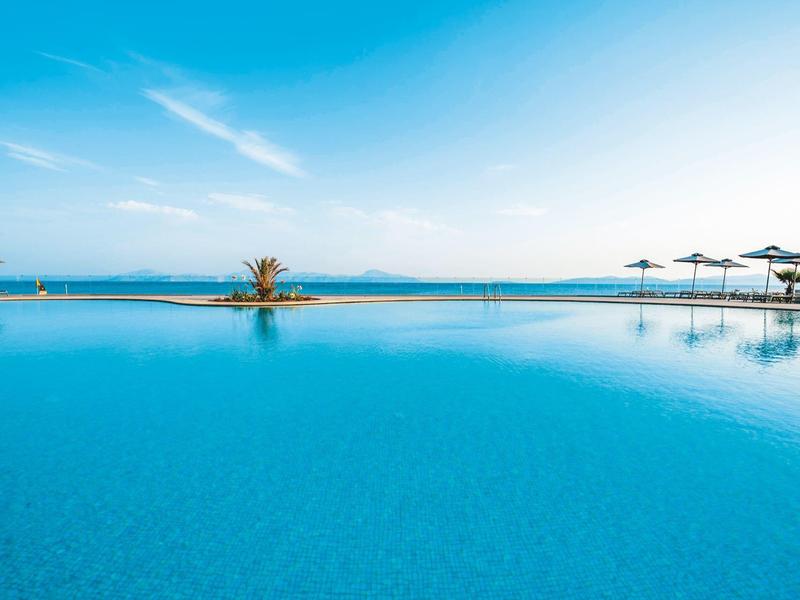 Vista di una piscina a sfioro con acqua azzurra limpida e ombrelloni lungo il bordo sotto un cielo soleggiato.