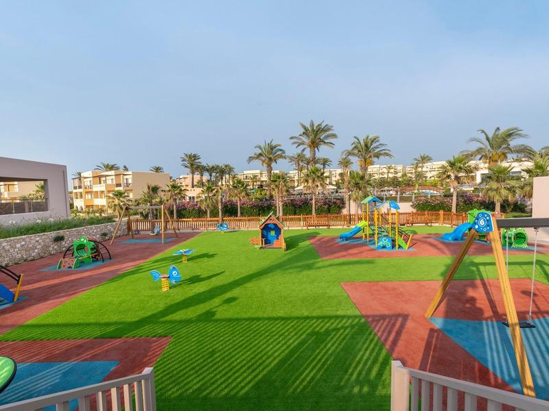 Area giochi colorata con strutture per arrampicarsi e scivoli, circondata da palme e edifici sotto un cielo sereno.