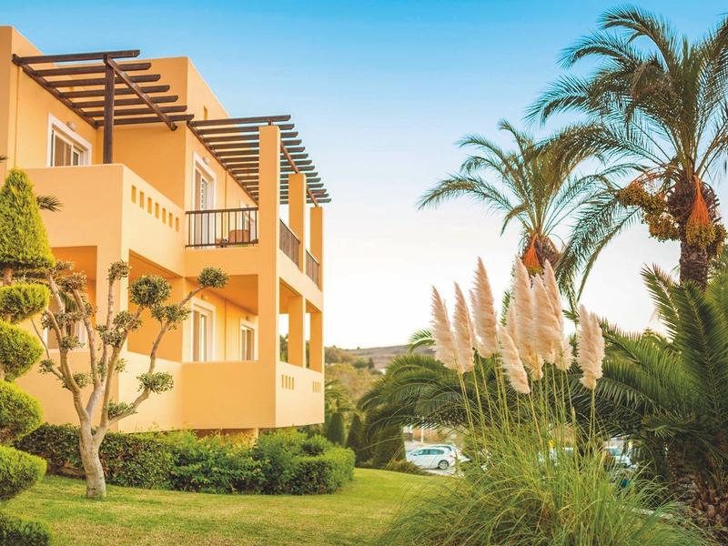 Edificio dell'hotel con balconi circondato da giardino curato e palme sotto un cielo limpido.