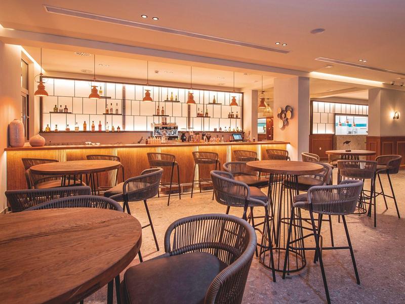 Area bar moderna in hotel con tavoli rotondi in legno, sedie accoglienti e bancone illuminato.