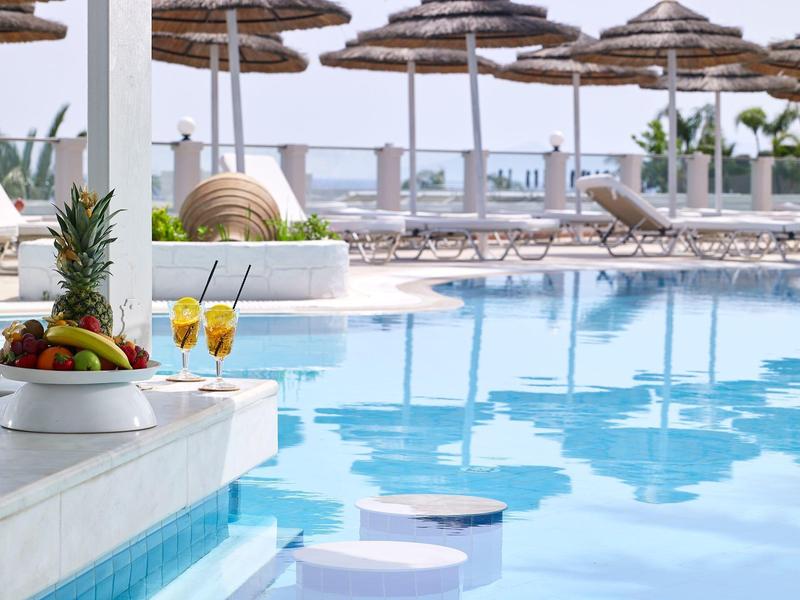 Piscina dell'hotel con sedili in acqua, ombrelloni e lettini sotto un cielo limpido.