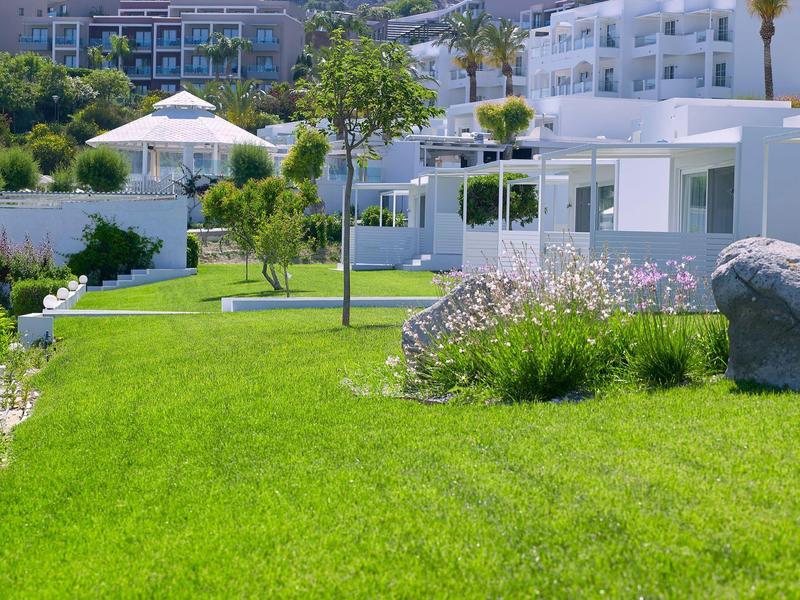 Giardino verde con alberi e fiori davanti a edifici bianchi dell'hotel in una giornata di sole.