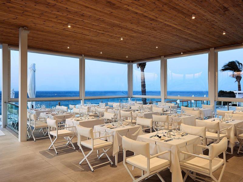 Area ristorante aperta sulla spiaggia con tavoli bianchi e vista mare al crepuscolo.