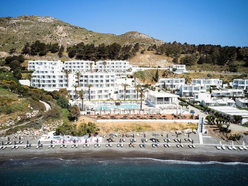 Grande hotel bianco con piscina su una collina vicino al mare con lettini sulla spiaggia.