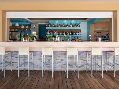 Area bar moderna in hotel con bancone bianco e nove sgabelli alti davanti a scaffali con bottiglie.