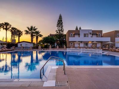 Grande piscina con lettini e palme davanti a moderni appartamenti al tramonto.