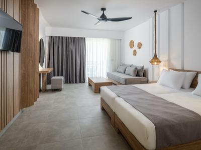 Camera d'hotel moderna con letto matrimoniale, divano, TV e ventilatore a soffitto in tonalità neutre.