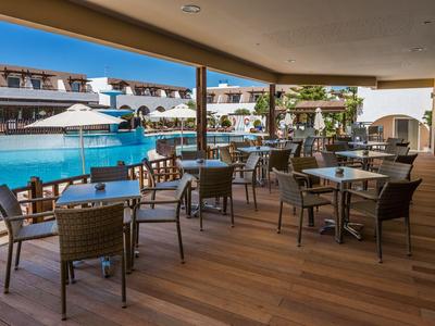 Terrazza coperta con tavoli e sedie accanto a una grande piscina dell'hotel sotto un cielo azzurro.