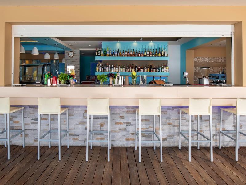 Area bar moderna in hotel con bancone bianco e nove sgabelli alti davanti a scaffali con bottiglie.