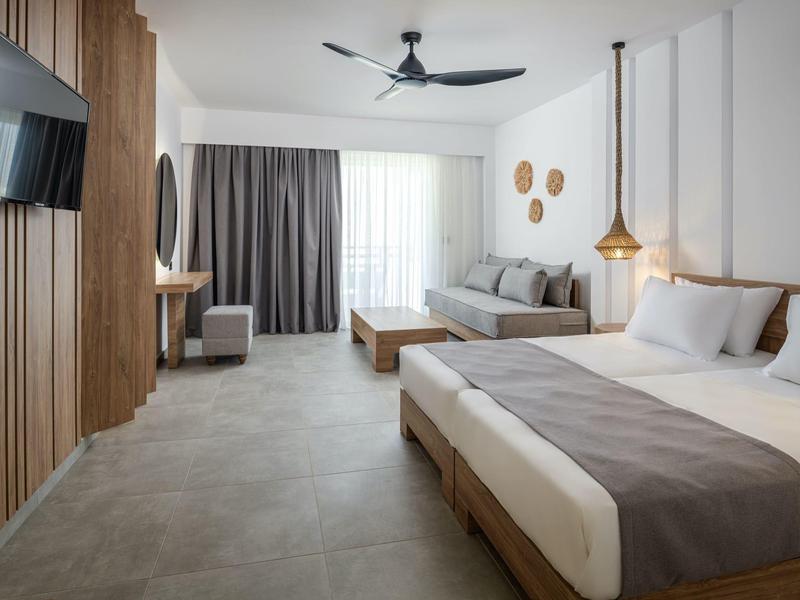 Camera d'hotel moderna con letto matrimoniale, divano, TV e ventilatore a soffitto in tonalità neutre.