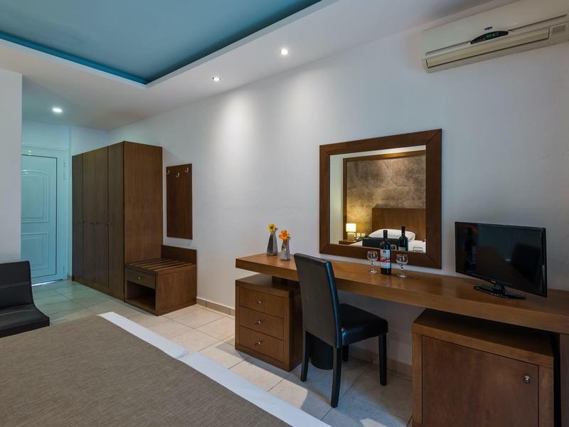 Camera d'hotel moderna con scrivania, sedia, TV e armadio in tonalità neutre.