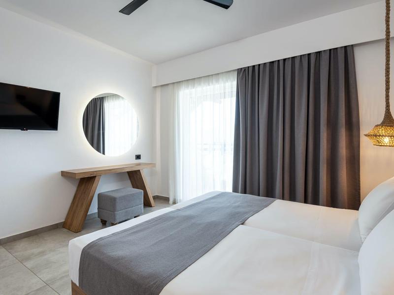 Camera d'hotel moderna con letto, TV, specchio, finestra con tende e lampada sospesa.