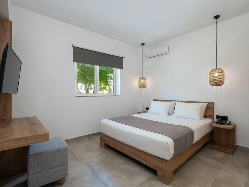 Camera d'hotel moderna con letto matrimoniale, TV, comodino e finestra con tendina grigia.