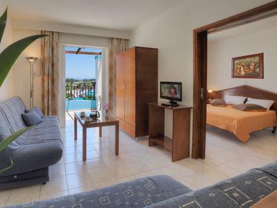 Helles Wohnzimmer mit blauer Couch, Holzmöbeln, offener Tür zum Schlafzimmer und Poolblick.