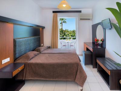 Ein helles Schlafzimmer mit Doppelbett, brauner Bettdecke, Schreibtisch, Fernseher und Balkon mit Aussicht.