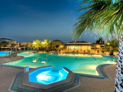 Beleuchteter Swimmingpool mit Whirlpool, Palmen und Lounge-Bereich bei Abenddämmerung.