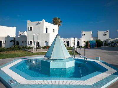 Kleiner Pool mit pyramidenförmiger Skulptur vor weißem Ferienhaus im sonnigen Ferienresort.