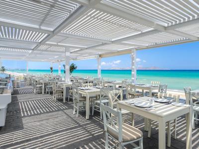 Hell eingerichtetes Strandrestaurant mit weißen Tischen und Stühlen unter einer Pergola mit Meerblick.