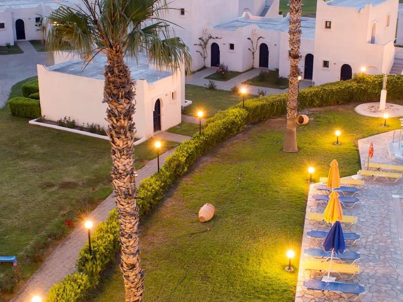 Mediterrane Hotelanlage mit weißen Gebäuden, Palmen und Pool bei Sonnenuntergang.