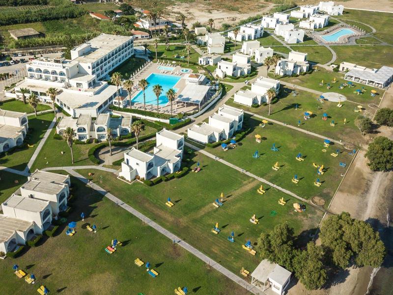 Luftaufnahme eines Strandresorts mit Gebäuden, Pool und Liegestühlen am Wasser.