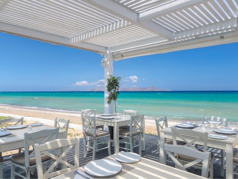 Ein offener Essbereich mit weißen Möbeln und Blick auf einen Strand und das Meer unter blauem Himmel.