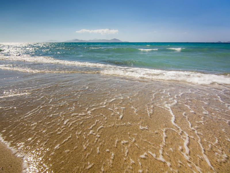 Klares Meerwasser und sanfte Wellen am sandigen Strand unter blauem Himmel mit Insel im Hintergrund.