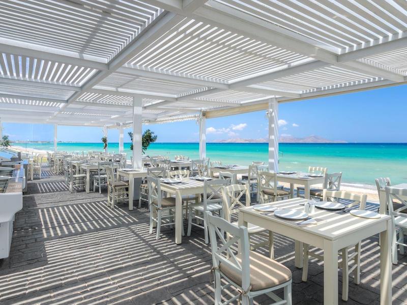 Hell eingerichtetes Strandrestaurant mit weißen Tischen und Stühlen unter einer Pergola mit Meerblick.