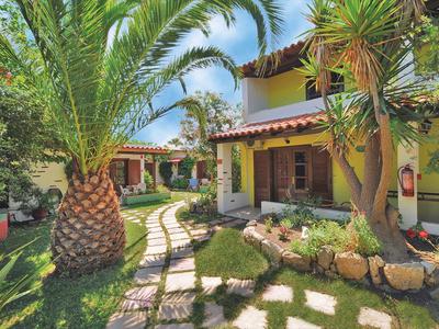 Giardino con palme e sentiero lastricato che porta a una casa gialla con tetti rossi sotto un cielo azzurro.