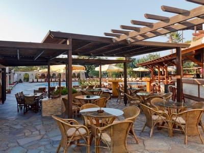 Zewnętrzny ogródek z wiklinowymi krzesłami i drewnianymi stołami pod pergolami na kamiennej podłodze.