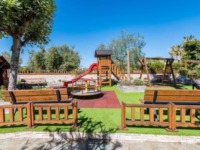 Słoneczny plac zabaw z drewnianymi ławkami, zjeżdżalnią, huśtawkami i zielonym trawnikiem pod jasnym niebem.
