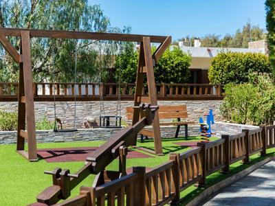 Plac zabaw na zewnątrz z huśtawką, wagą, ławkami, trawą i drewnianym płotem pod czystym niebem.