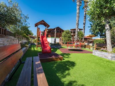 Plac zabaw z zjeżdżalnią i huśtawką otoczony ławkami i drzewami pod bezchmurnym niebem.