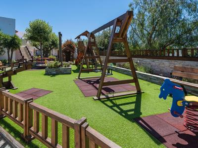 Plac zabaw na zewnątrz z huśtawkami i sprężynowymi zabawkami na sztucznej trawie, otoczony drewnianymi ławkami i ogrodzeniem.