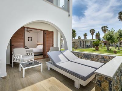 Balcone con lettini e tavolo, vista sul giardino con palme e edificio bianco.